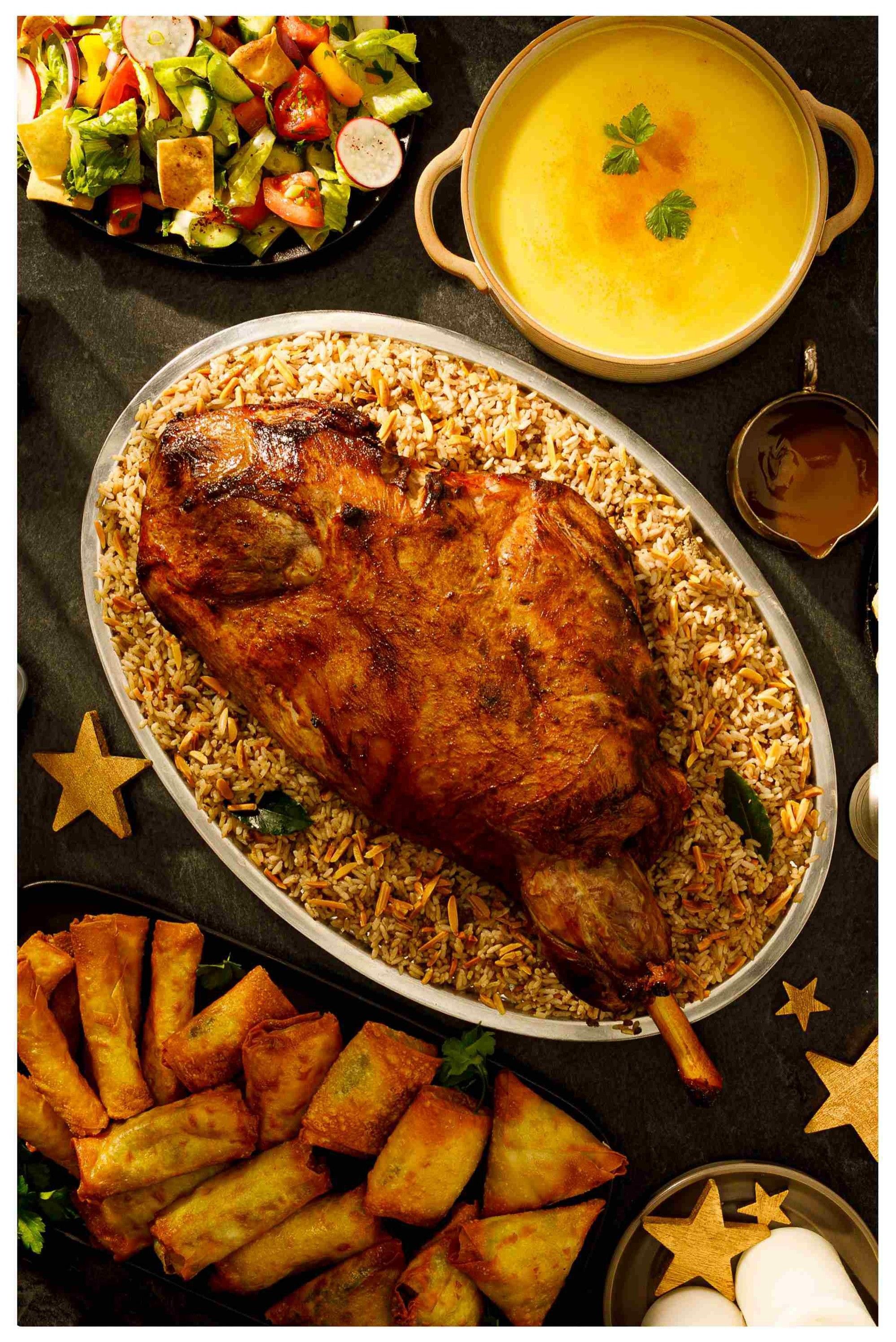 Adonis weekly flyer specials page 17 features a festive spread including a whole roasted lamb served over a bed of rice with slivered almonds. Accompanying dishes include a fresh salad with lettuce, tomatoes, cucumbers, and radishes, a bowl of creamy yellow soup, and a platter of golden-brown fried spring rolls or samosas. Decorative wooden stars and a small dish of sauce are also visible, suggesting a celebratory meal.